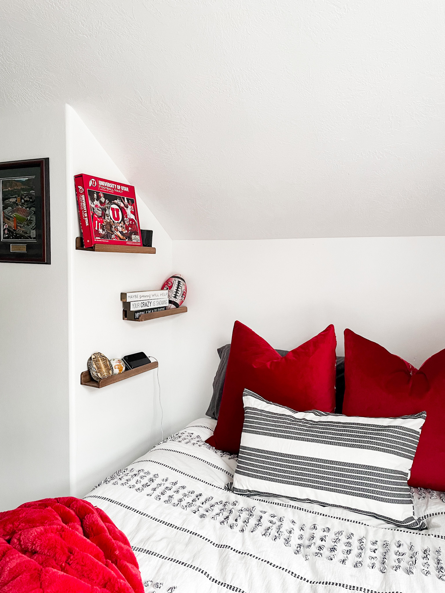 three shelves with book phone and football on them, red pillows and red blanket, white wall with shelves